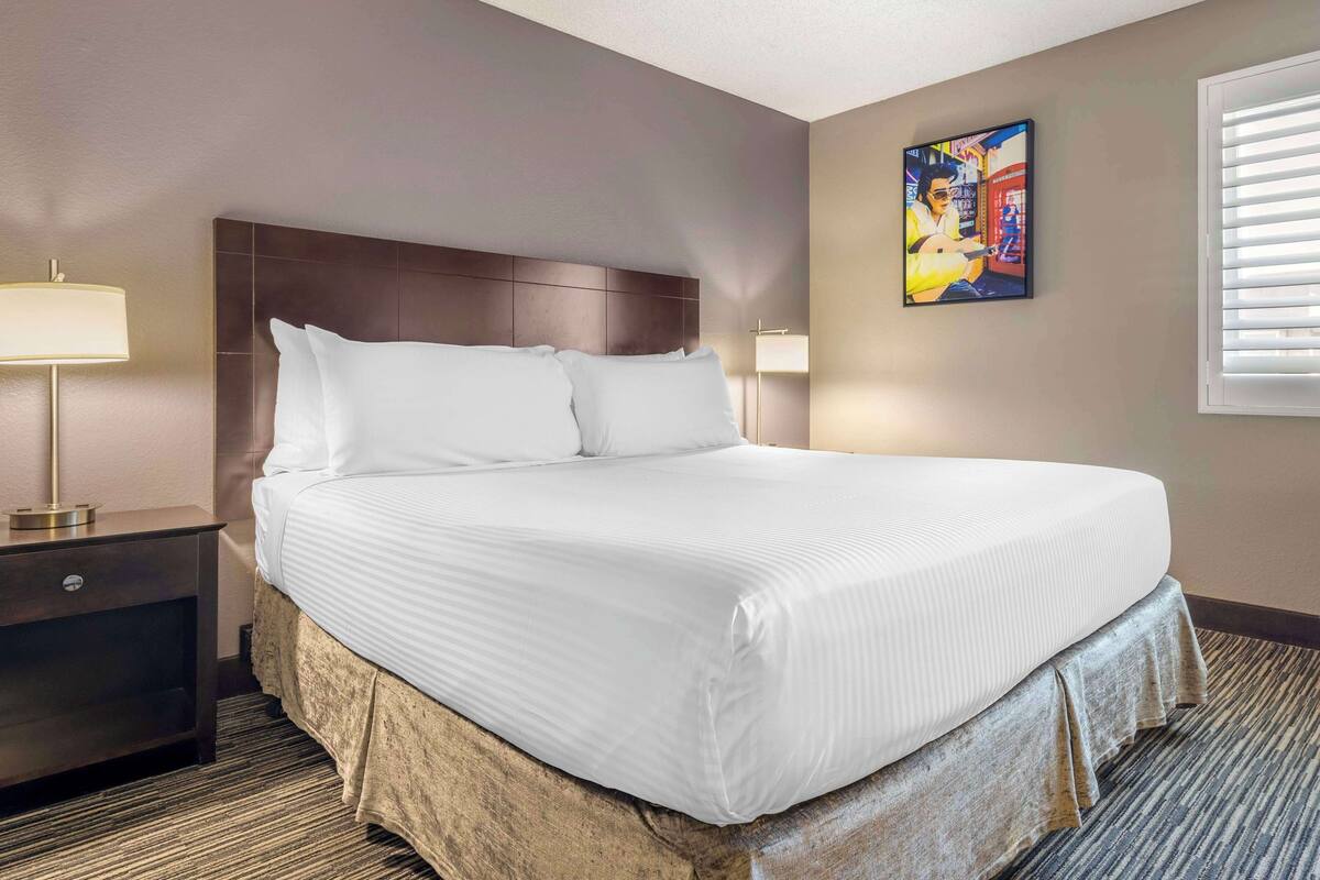 A hotel room with a large bed, a nightstand with a lamp, a wall-mounted artwork, and a window with blinds.