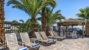 A sunlit outdoor seating area with lounge chairs and a thatched umbrella, surrounded by palm trees and a stone wall. - Best Western McCarran Inn (Las Vegas)