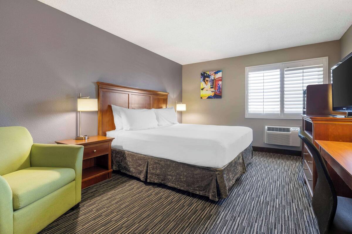 A hotel room with a large bed, a desk with a television, a chair, and a window with blinds.