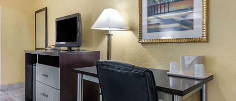 A hotel room with a desk, a chair, a television, a lamp, a framed painting, and a minibar.