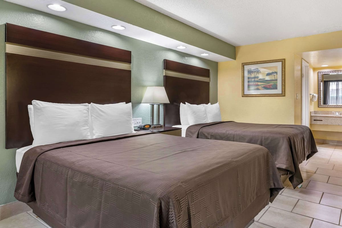 A hotel room with two beds, a nightstand with a lamp, a painting on the wall, and a bathroom visible through an open door.