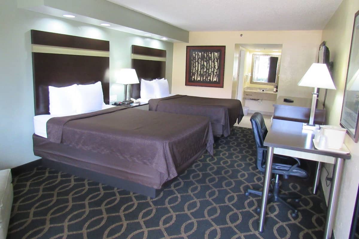 A hotel room with two beds, a desk, a chair, a desk lamp, a mirror, and a patterned carpet.