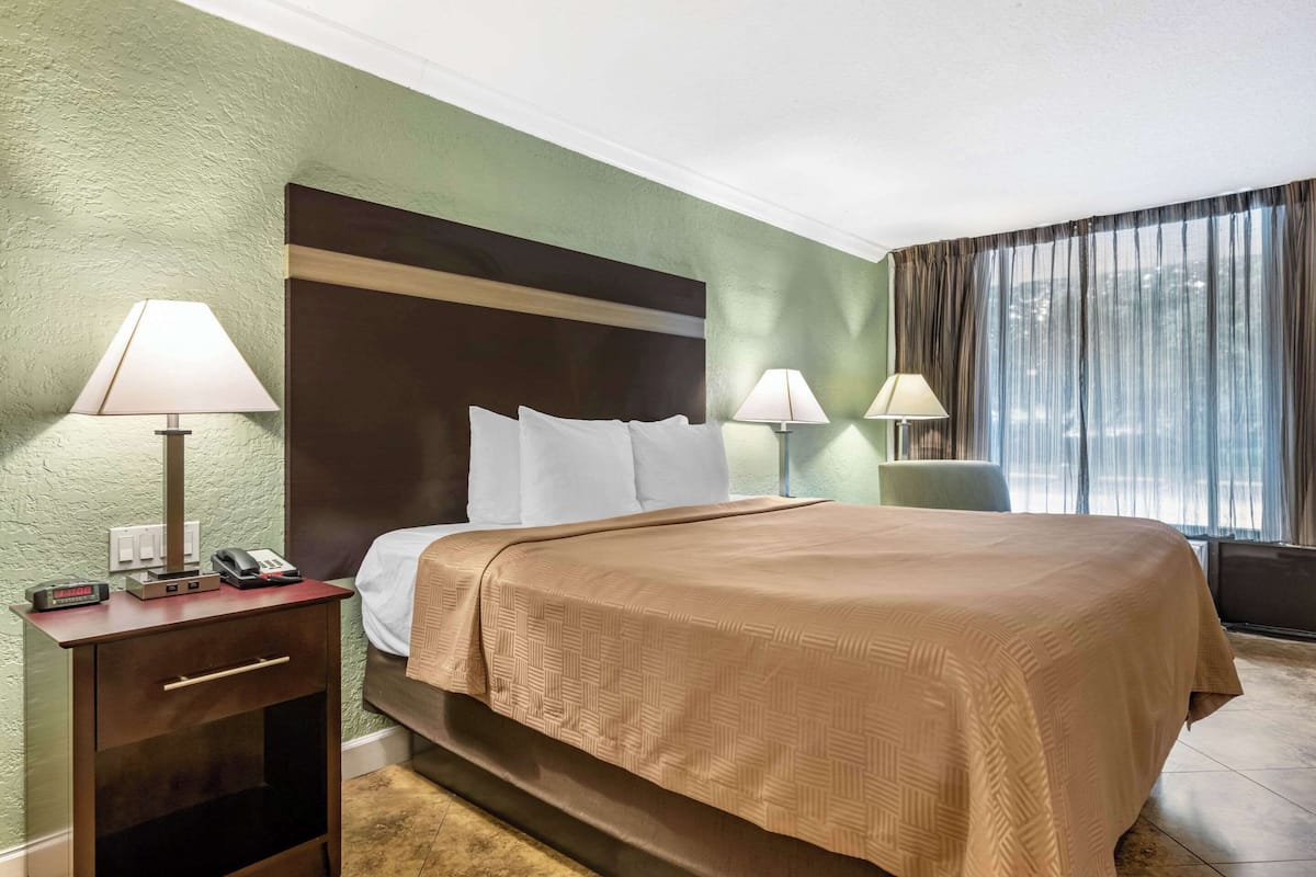 A hotel room with a bed, two bedside lamps, a nightstand with a phone and a clock, and a window with curtains.