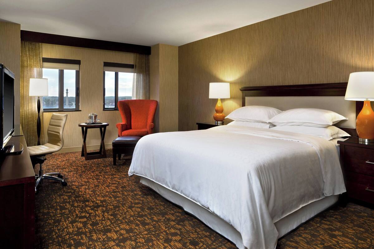 A hotel room with a large bed, two bedside lamps, a desk with a chair, a television, and a view of the city through the windows.