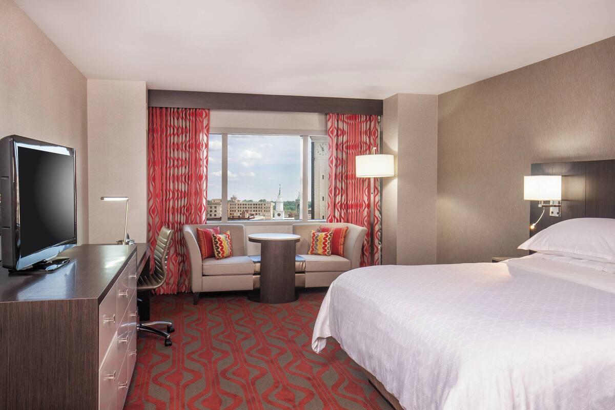 A hotel room with a bed, a desk with a TV, a sofa, a small table, and a view of the city through large windows.