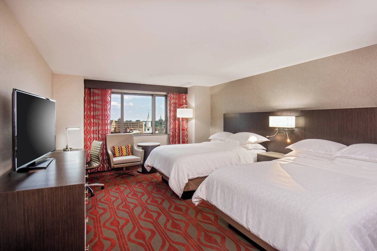 A hotel room with two beds, a desk with a TV, a chair, a window with curtains, and a view of the city.