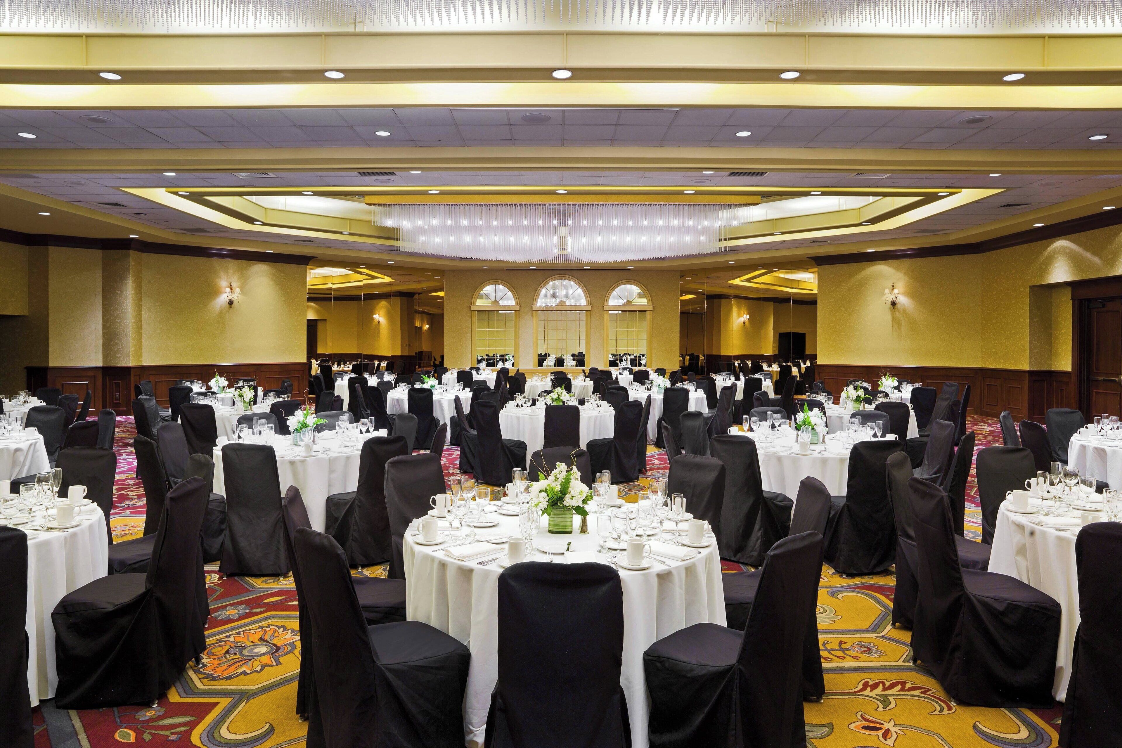 A large banquet hall with round tables set for dining, black chairs, and chandeliers.