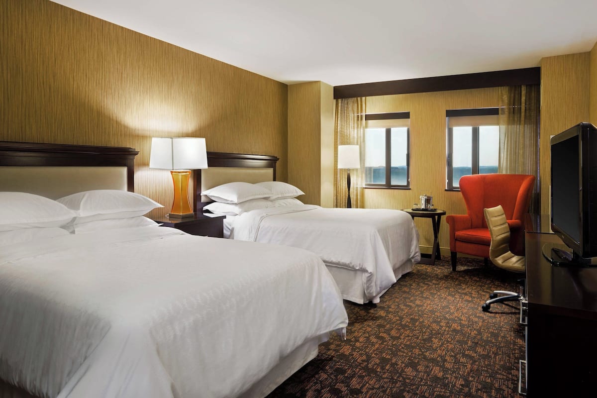 A hotel room with two beds, a TV, a chair, and a small table with lamps.