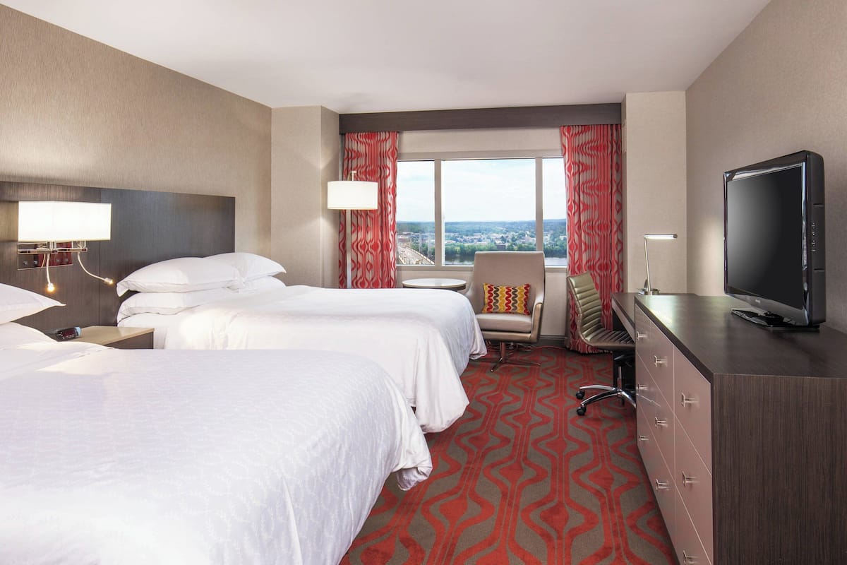 A hotel room with two beds, a TV, a desk, and a view of the city.