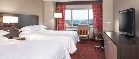 A hotel room with two beds, a TV, a desk, and a view of the city.