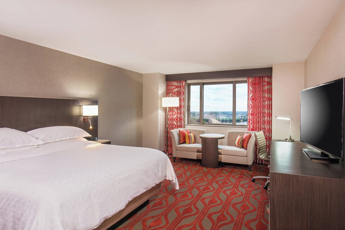A hotel room with a large bed, a desk with a TV, a sofa, and a view of the city.