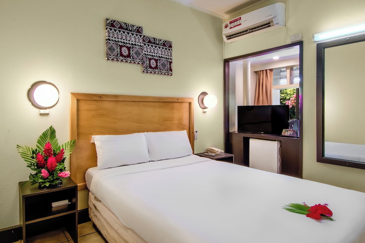 A hotel room with a bed, bedside tables, a TV, a mirror, and an air conditioning unit.