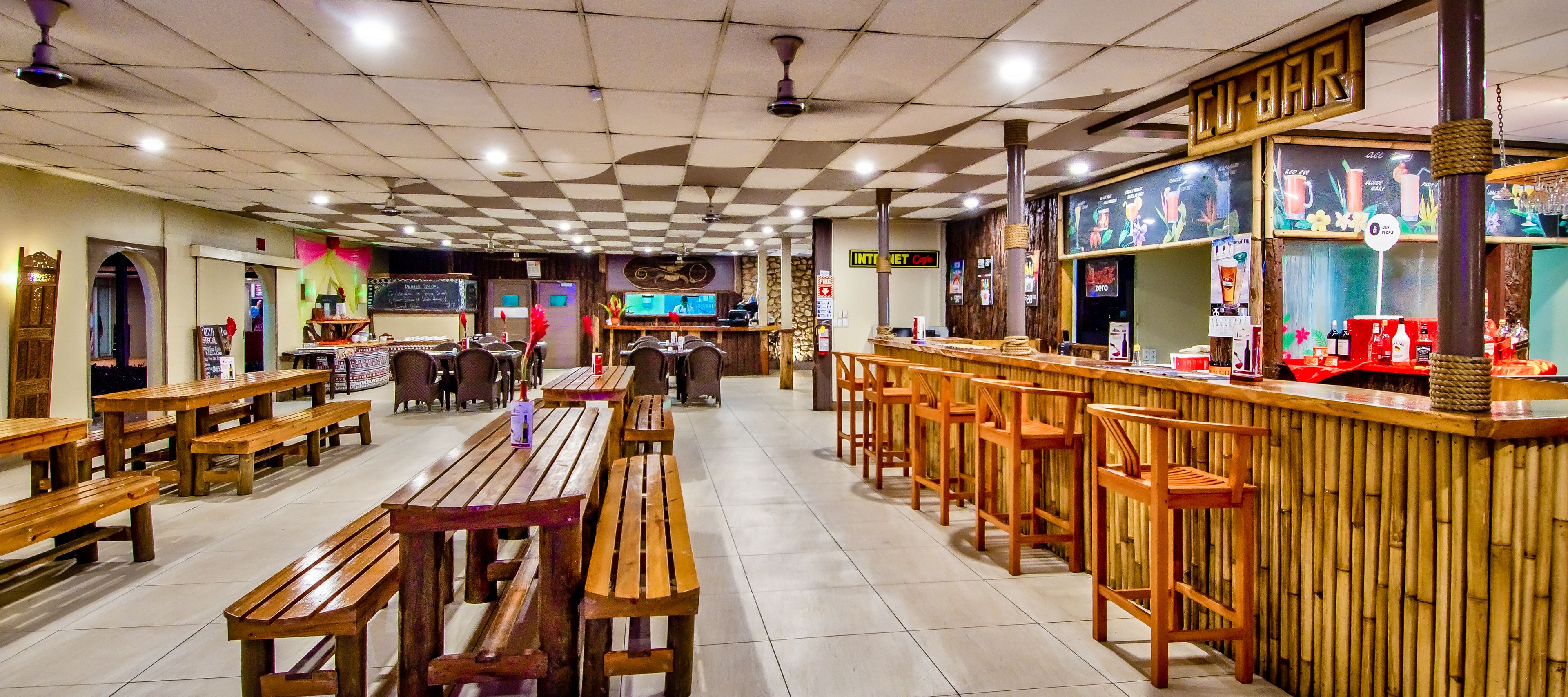 Interior de un restaurante con mesas y bancos de madera, una barra con menú de bebidas y un letrero que indica una cafetería con internet.