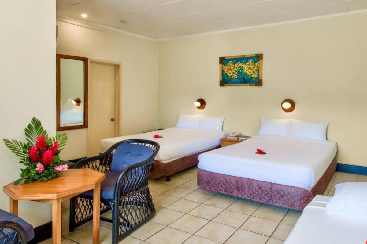 A hotel room with two beds, a small table with a chair, a decorative plant, and a painting on the wall.