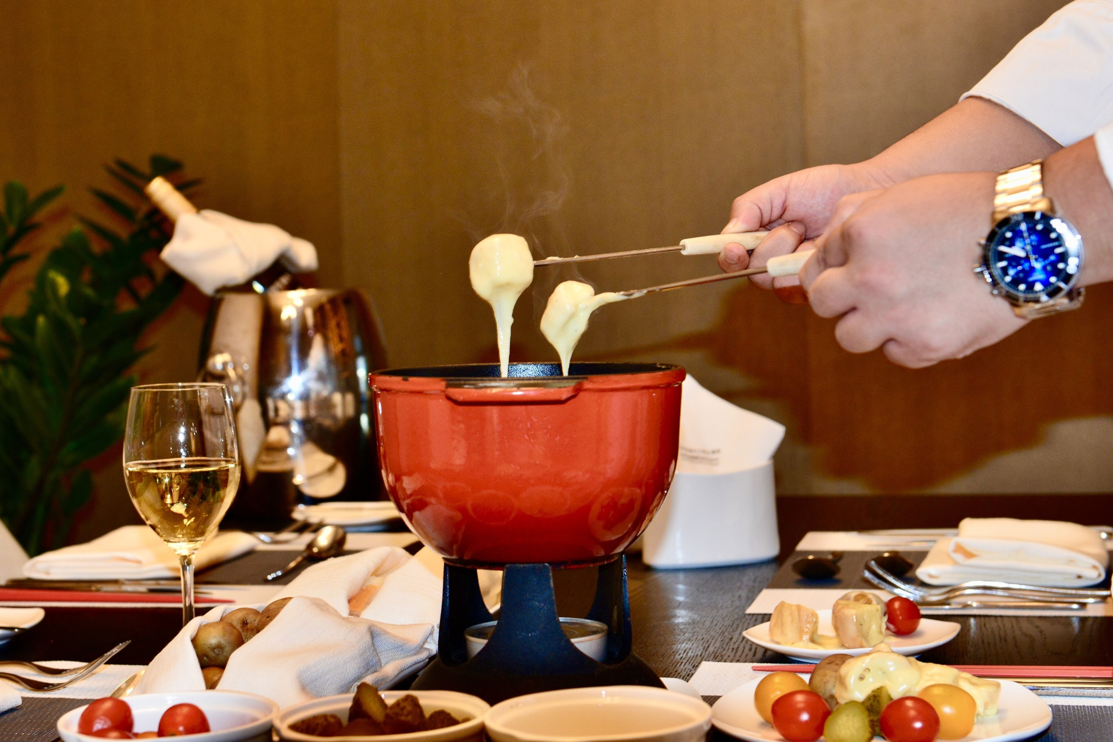 A dining table with a fondue pot, a glass of white wine, and various food items.