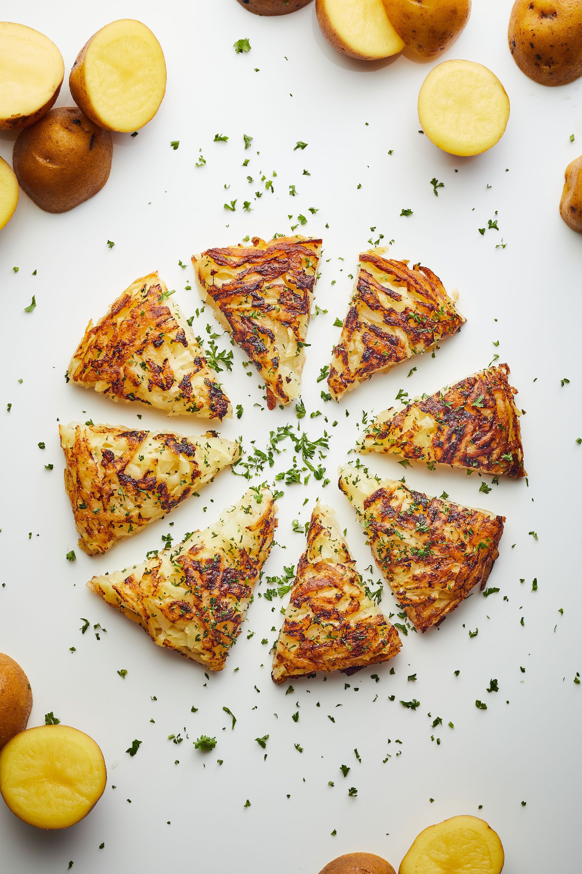 A white plate with golden-brown potato wedges arranged in a circular pattern, surrounded by whole and halved potatoes, garnished with green herbs.