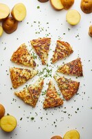 A white plate with golden-brown potato wedges arranged in a circular pattern, surrounded by whole and halved potatoes, garnished with green herbs.