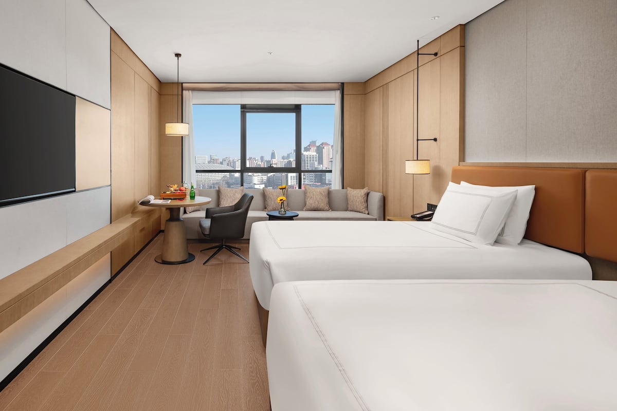 Ein Hotelzimmer mit zwei Betten, einem Fernseher, einem Schreibtisch mit Stuhl, einer Sitzecke und Blick auf die Stadt.