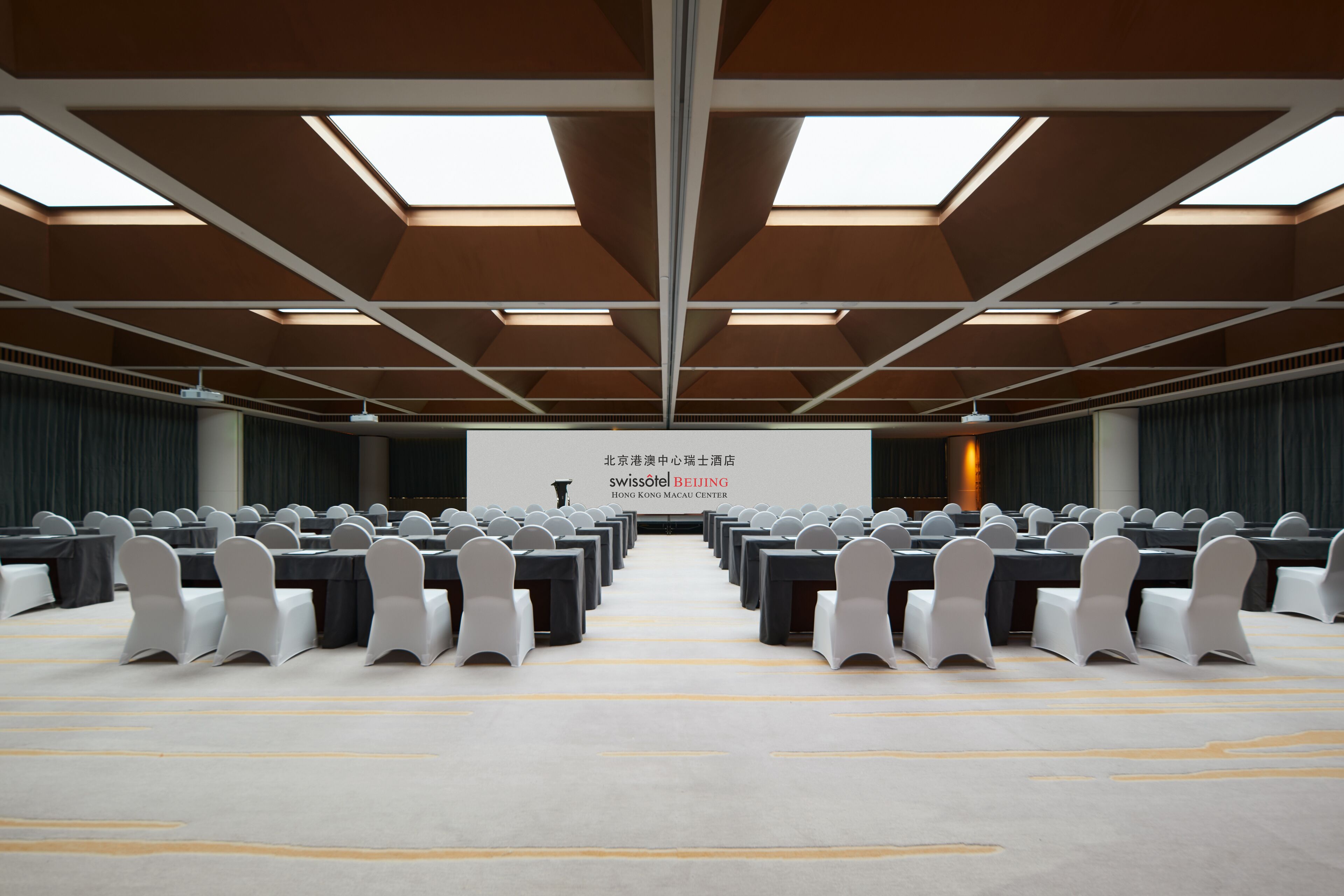 Un salón de conferencias con filas de mesas y sillas, una pantalla grande al frente y tragaluces en el techo.