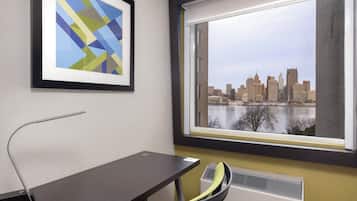 Una habitación con escritorio, silla, una obra de arte enmarcada y ventana con vista a la ciudad.