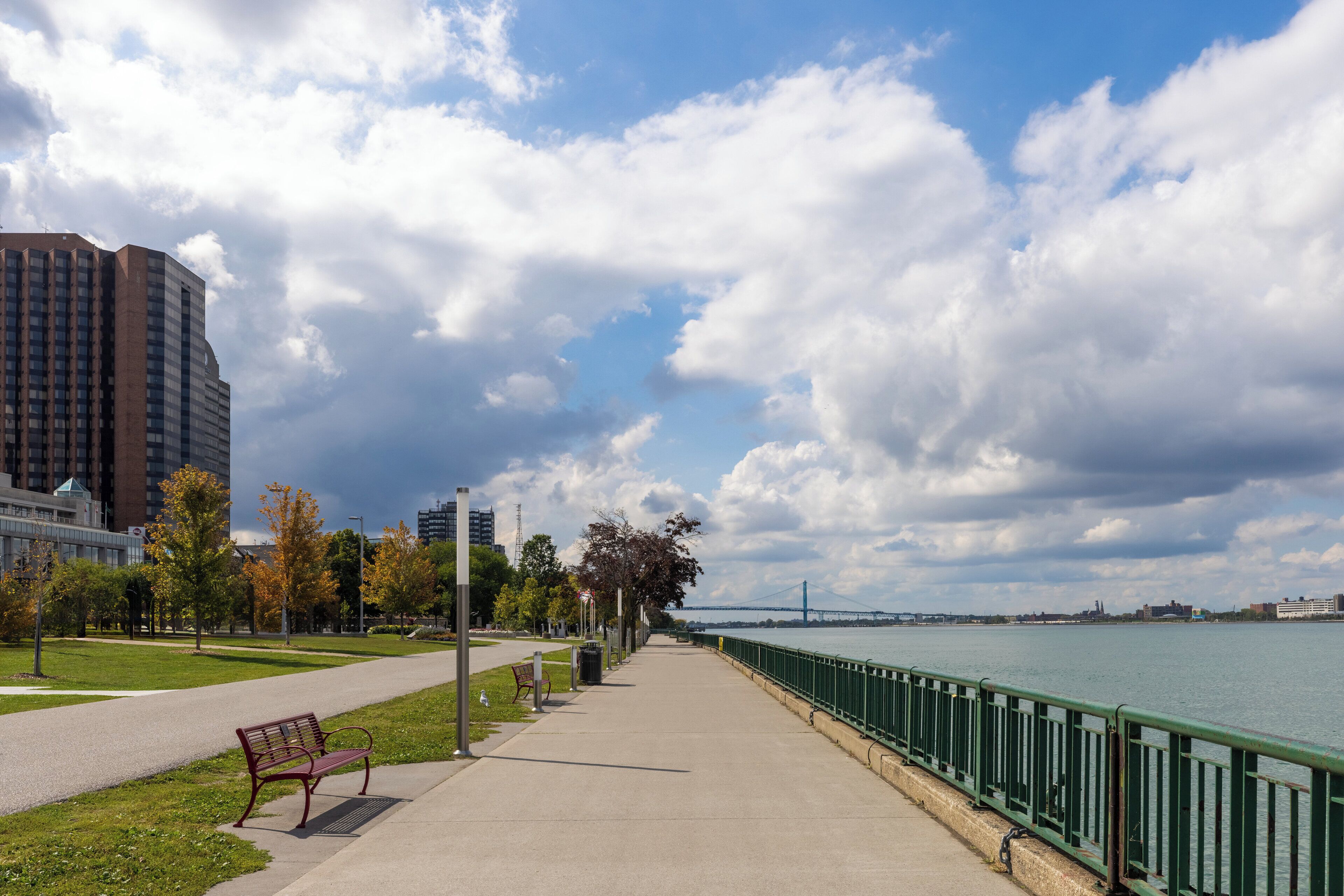 A riverside walkway with a bench, a metal railing, and a view of a bridge in the distance.