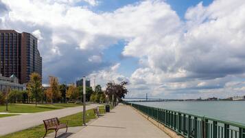 A riverside walkway with a bench, a metal railing, and a view of a bridge in the distance.