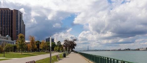 A riverside walkway with a bench, a metal railing, and a view of a bridge in the distance.
