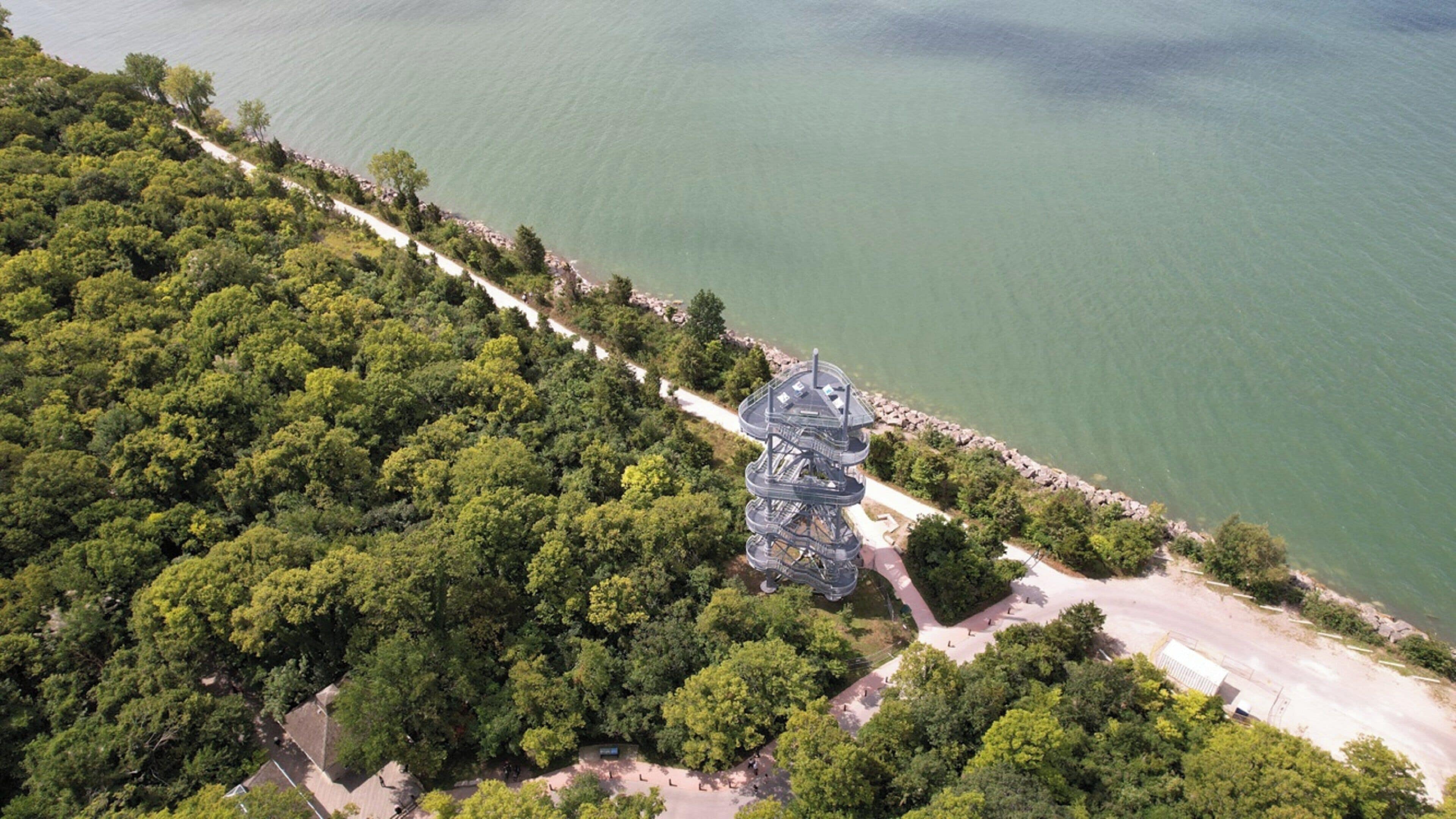 An aerial view of a coastal area with dense greenery, a winding road, and a large, multi-tiered structure near the shore.