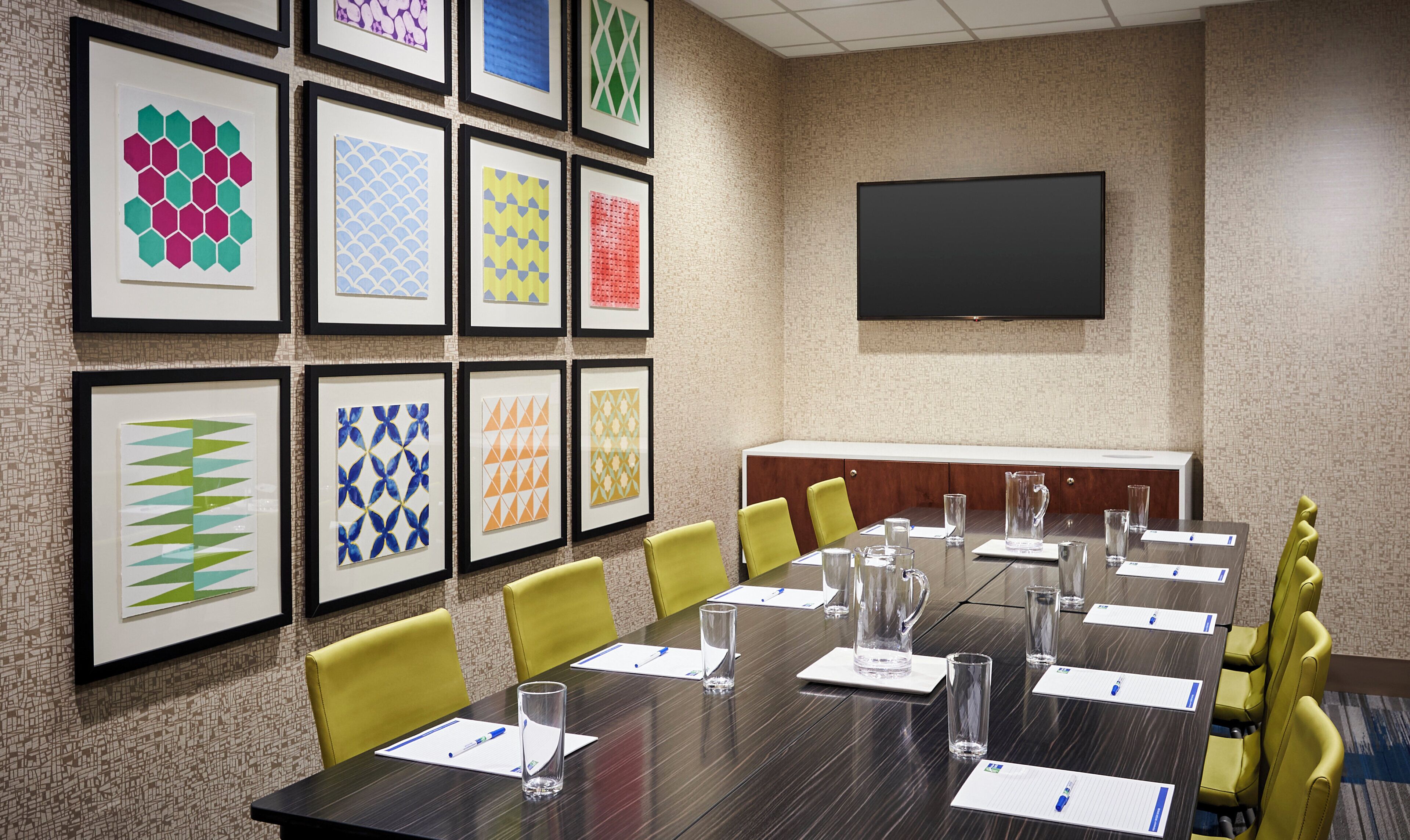A conference room with a long table, yellow chairs, framed artwork on the wall, and a flat-screen TV.
