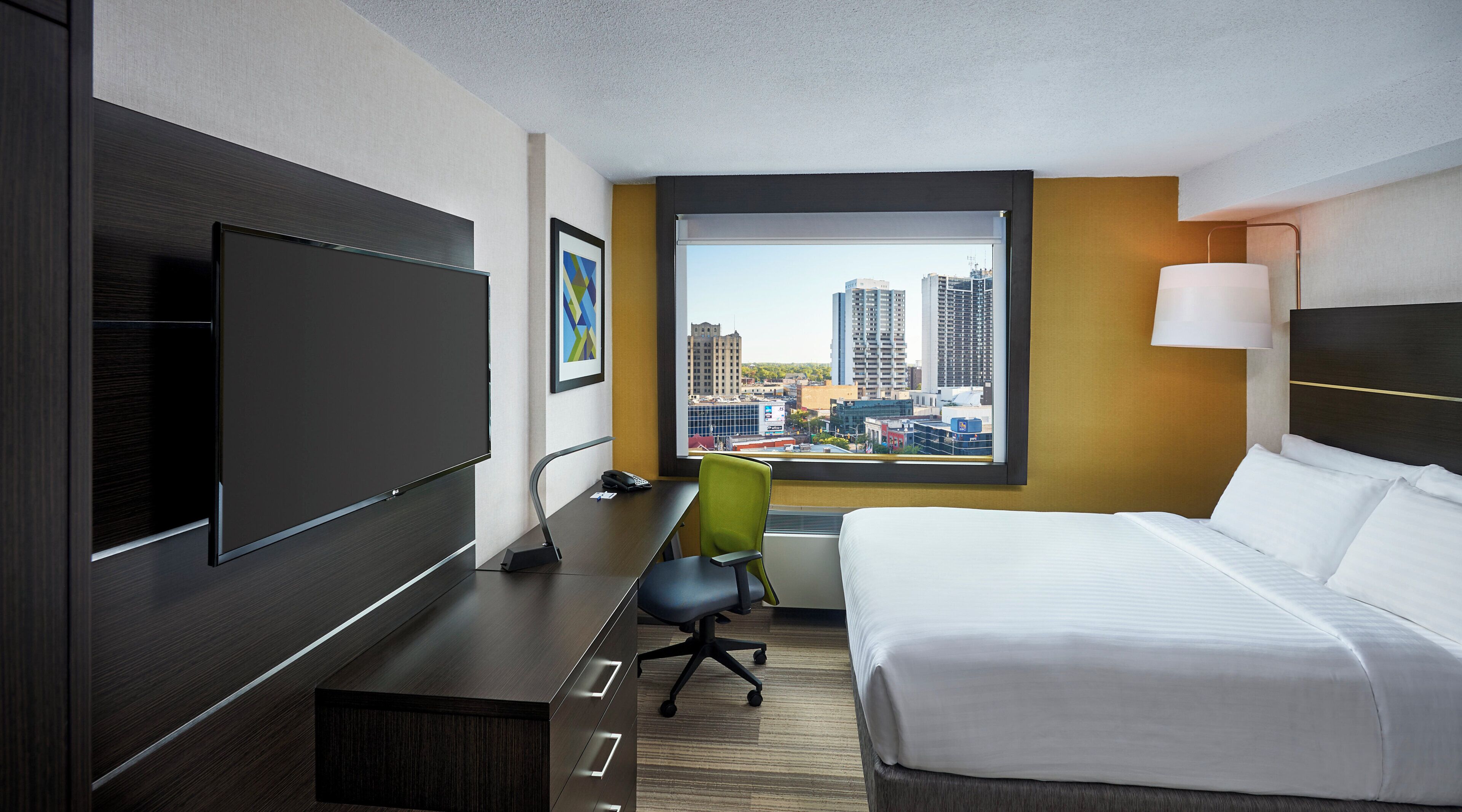 A hotel room with a bed, desk, chair, TV, and a window with a city view.
