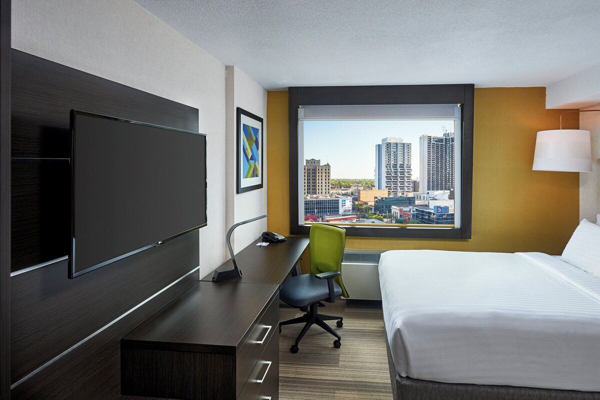 A hotel room with a bed, a desk, a chair, a TV, a window with a city view, and a lamp.