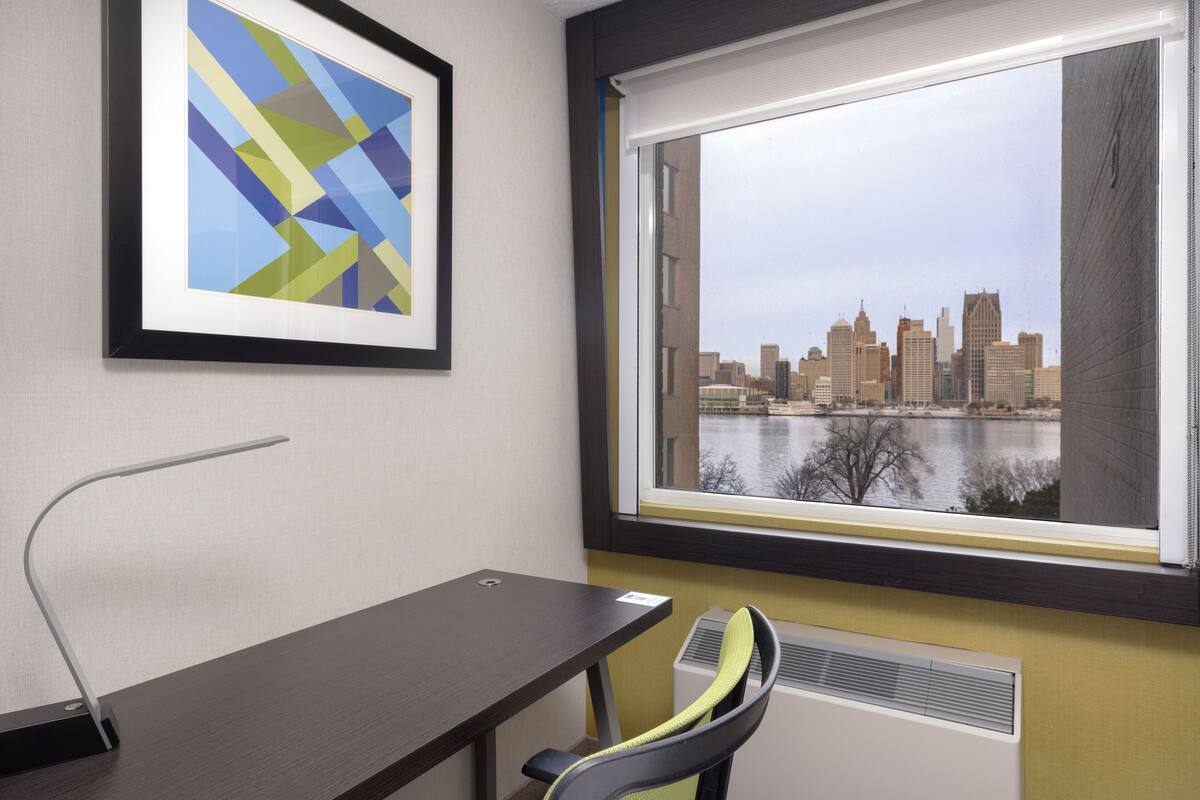 Une chambre d’hôtel équipée d’un bureau, d’une chaise, d’œuvres d’art encadrées et d’une fenêtre donnant sur le paysage urbain.