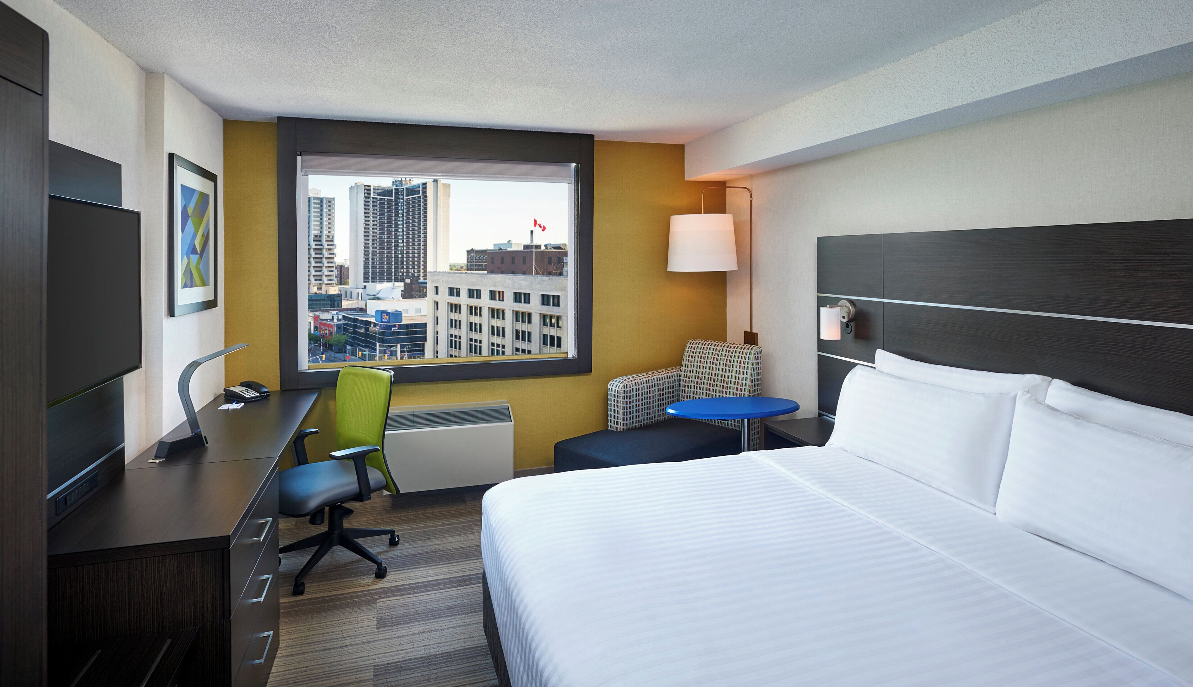 Habitación de hotel con cama, escritorio, silla y vista a un paisaje urbano a través de una ventana.