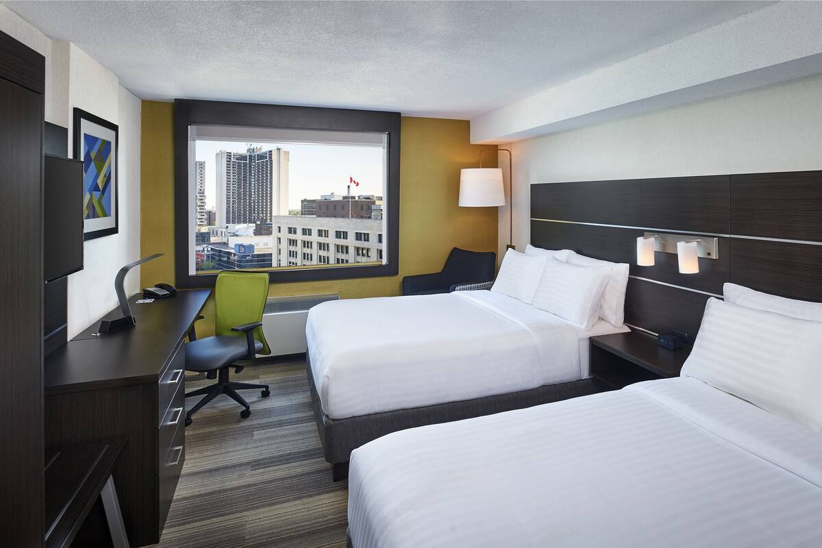 Une chambre d’hôtel avec deux lits, un bureau, une chaise, une fenêtre donnant sur des immeubles et un téléviseur fixé au mur.