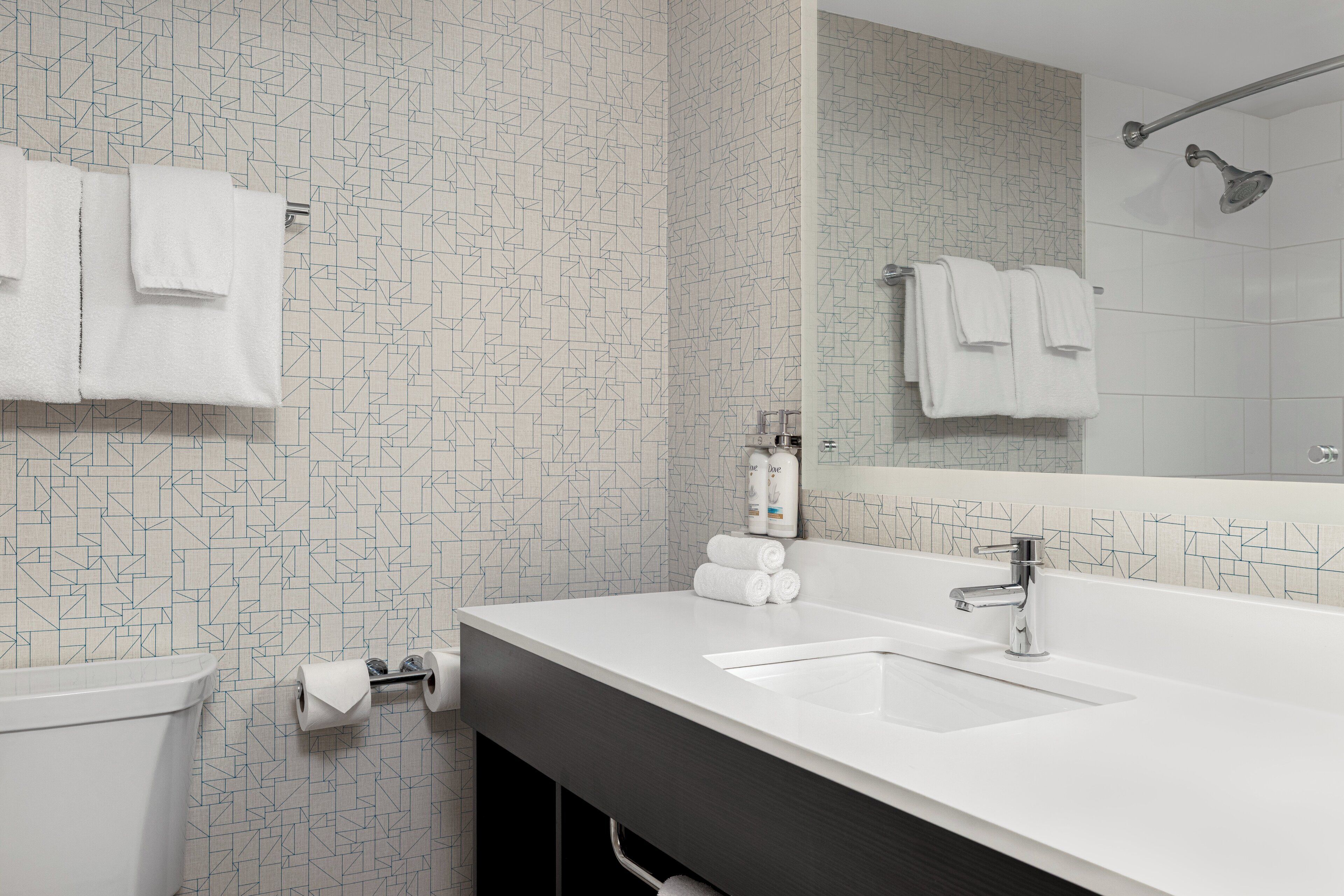 Une salle de bain moderne avec un lavabo blanc, un miroir et une douche. Des serviettes sont accrochées au mur, et il y a un porte-rouleau de papier toilette.