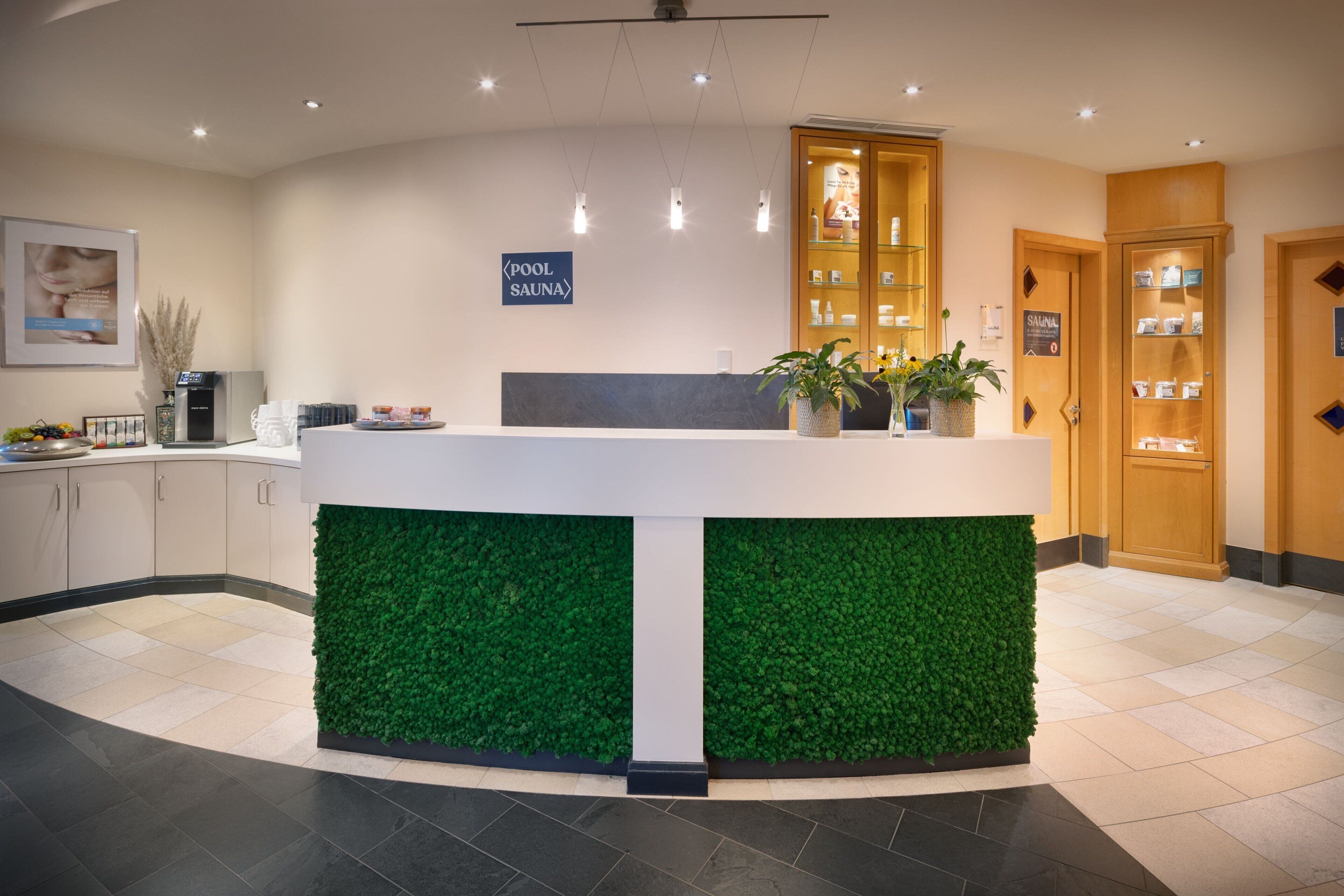 Zona de recepción con pared verde, un cartel que indica la piscina y sauna, y armarios de madera con productos.