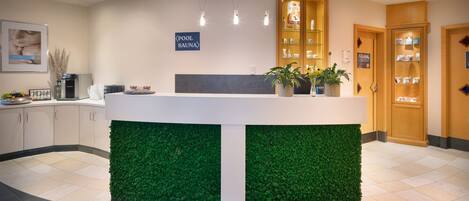 Zona de recepción con pared verde, un cartel que indica la piscina y sauna, y armarios de madera con productos.