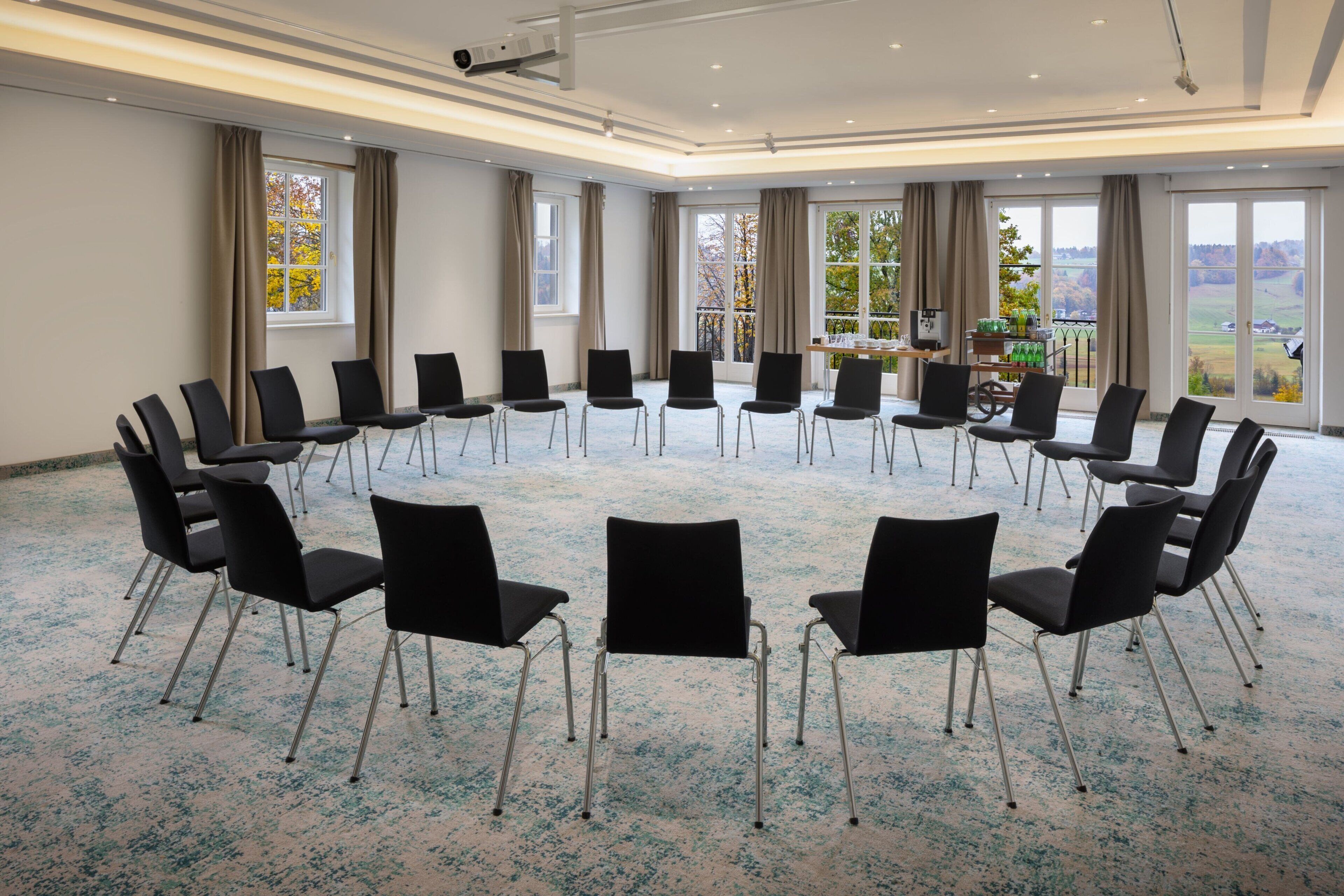 Une salle de conférence avec des chaises disposées en cercle, de grandes fenêtres avec des rideaux et une vue sur l’extérieur.
