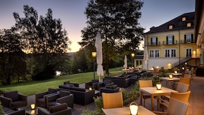 An outdoor seating area with wicker furniture and tables, surrounded by greenery and a building with balconies.
