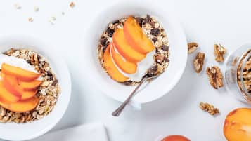 Two bowls of granola with sliced peaches and a spoon, surrounded by walnuts and a whole peach on a white surface.