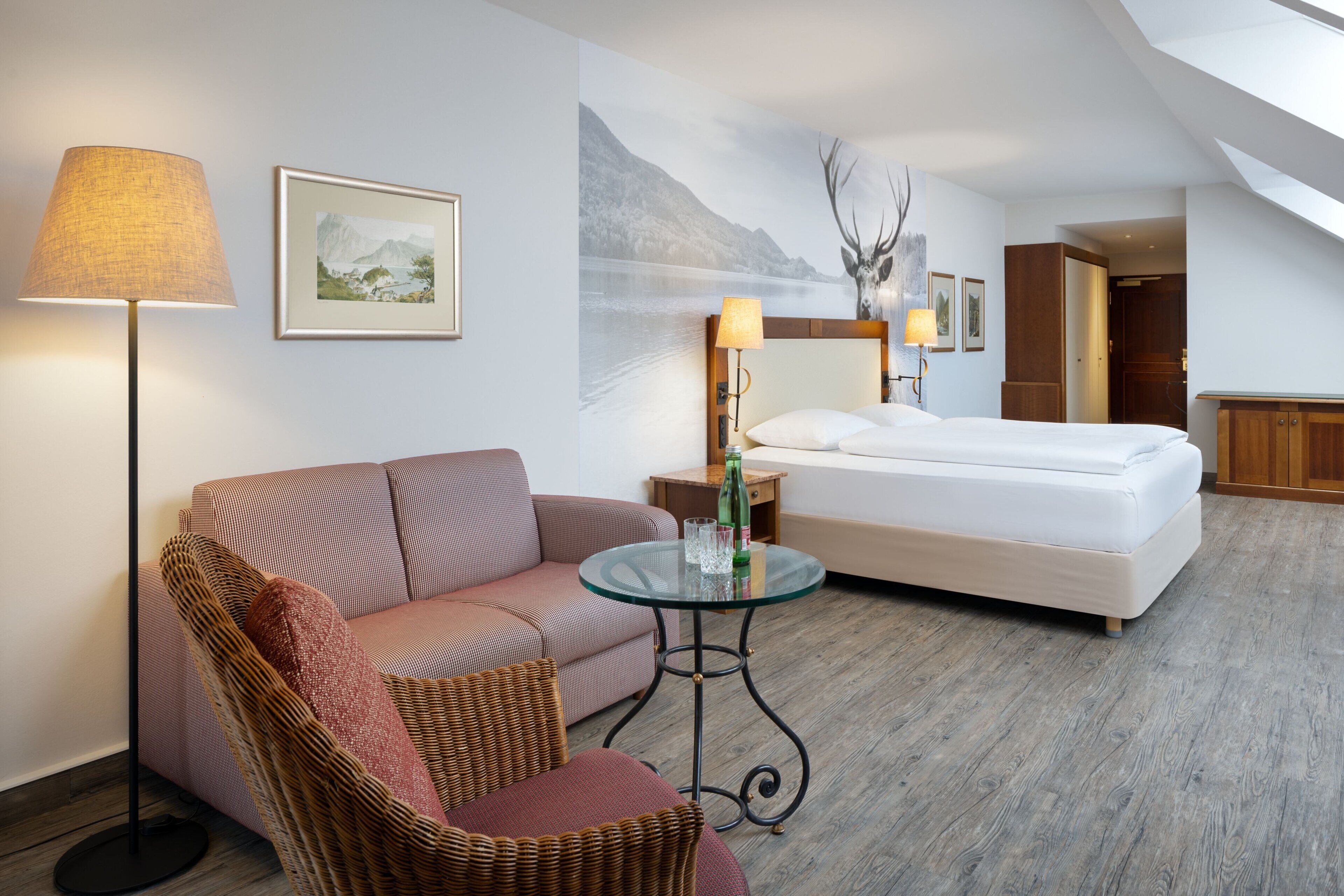 Una habitación de hotel con cama, sofá, sillón, una mesita y una lámpara.
