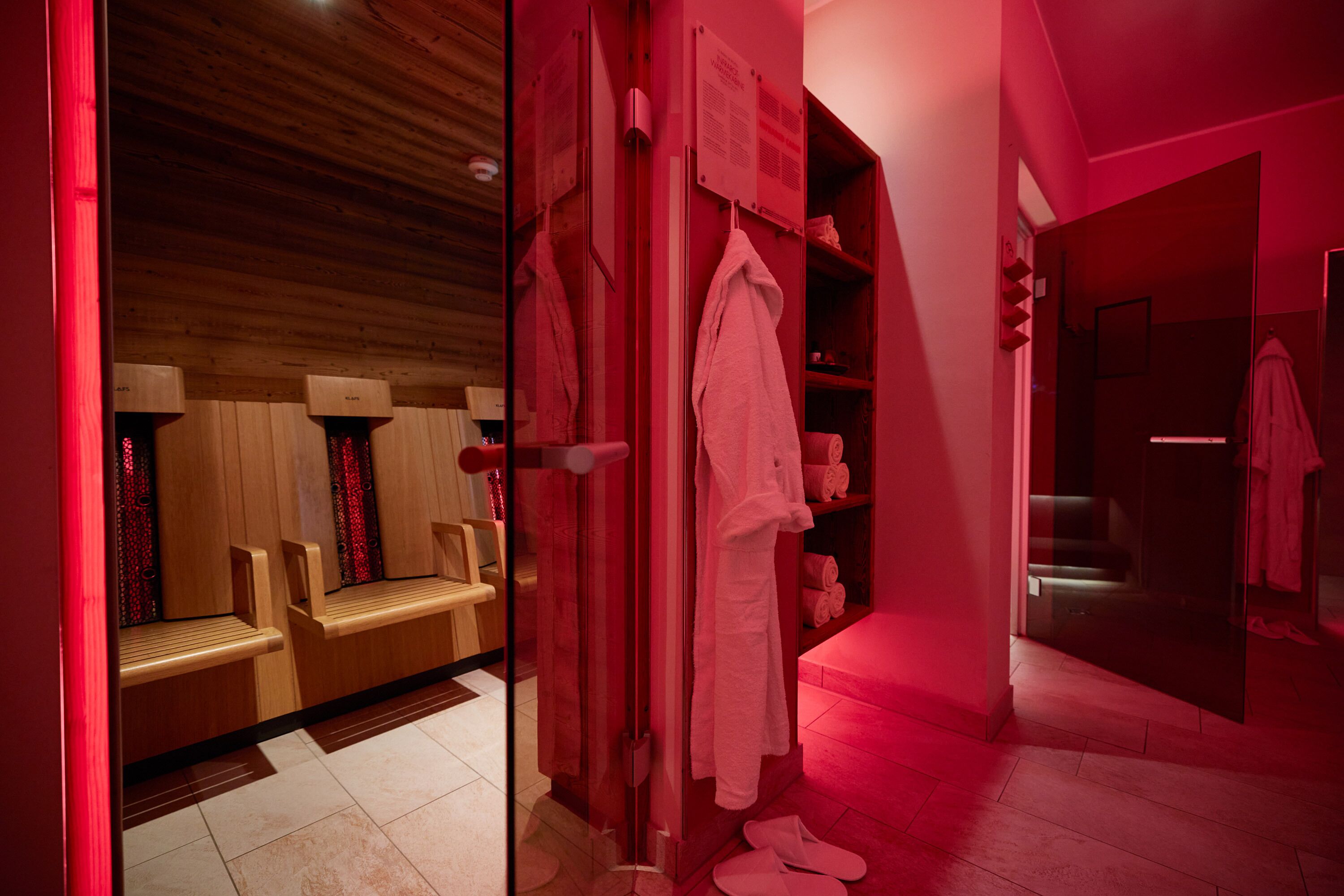 A sauna room with wooden benches and a glass door leading to a shower area with a red-lit ambiance.