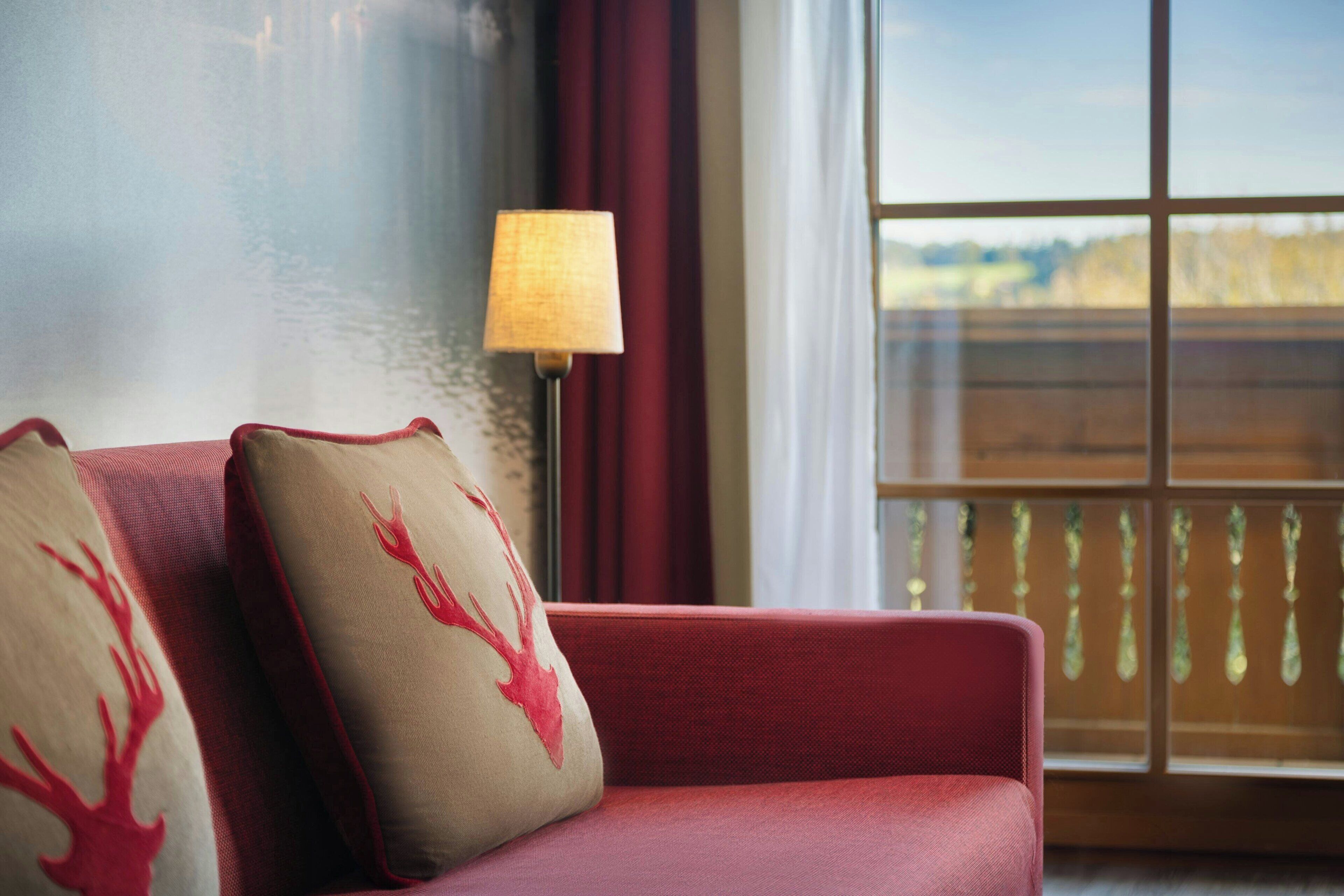 A red sofa with two decorative pillows, a floor lamp, and a view of a wooden deck through a window.
