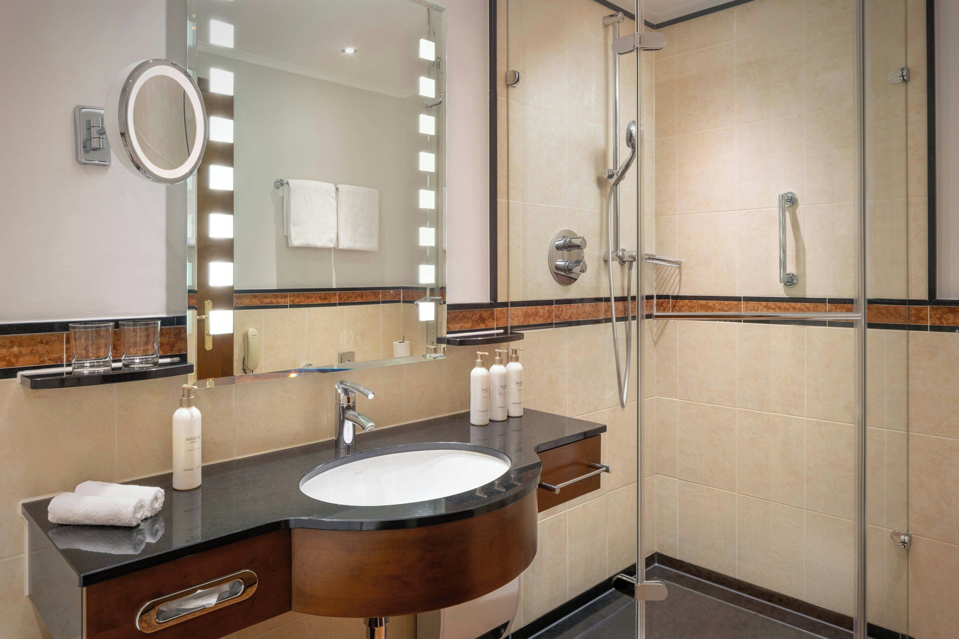 Un baño moderno con un lavabo redondo, un espejo con luces, una cabina de ducha de vidrio y un perchero para toallas.