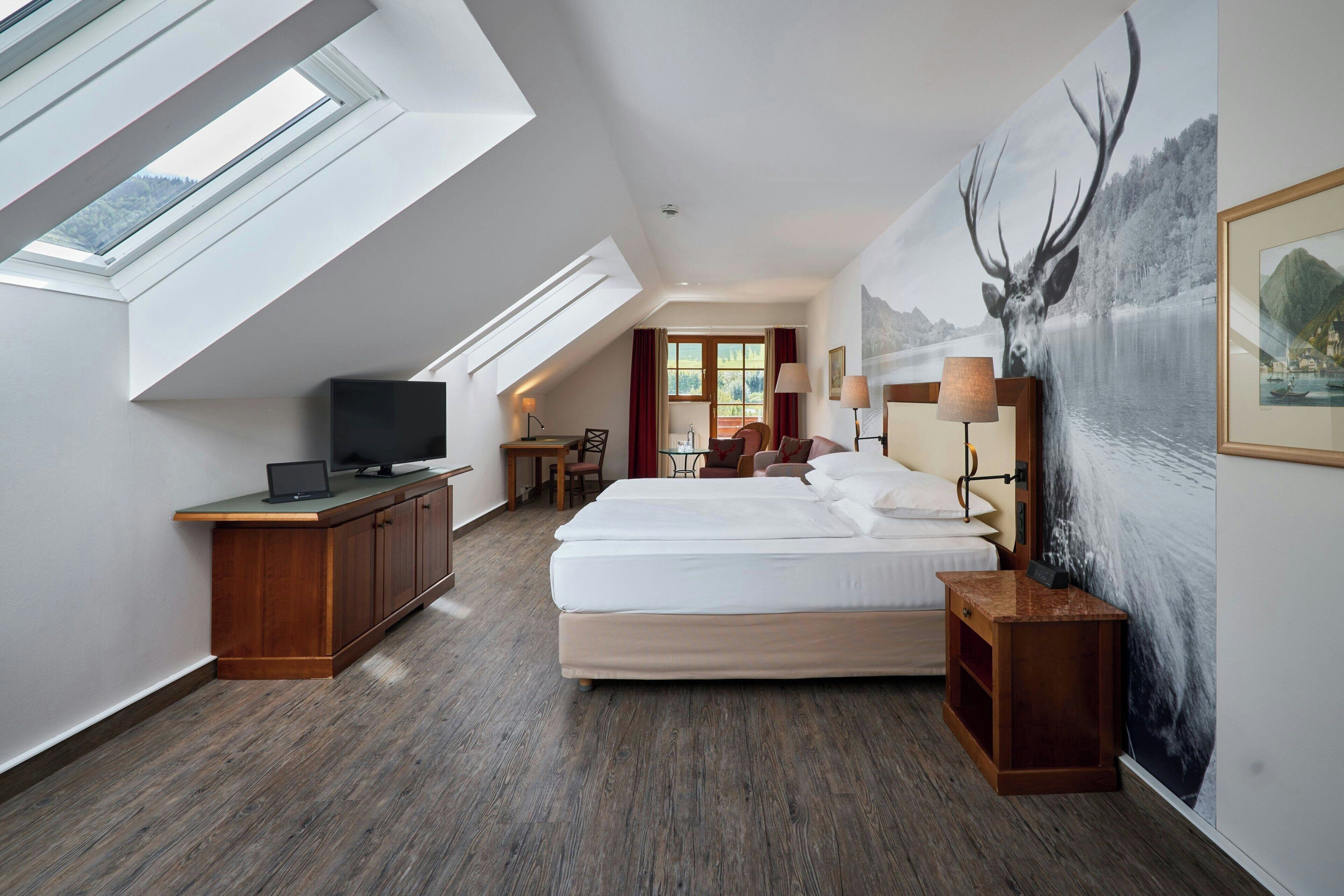 A modern hotel room with a large bed, a desk with a laptop, a TV, and a painting of a deer.
