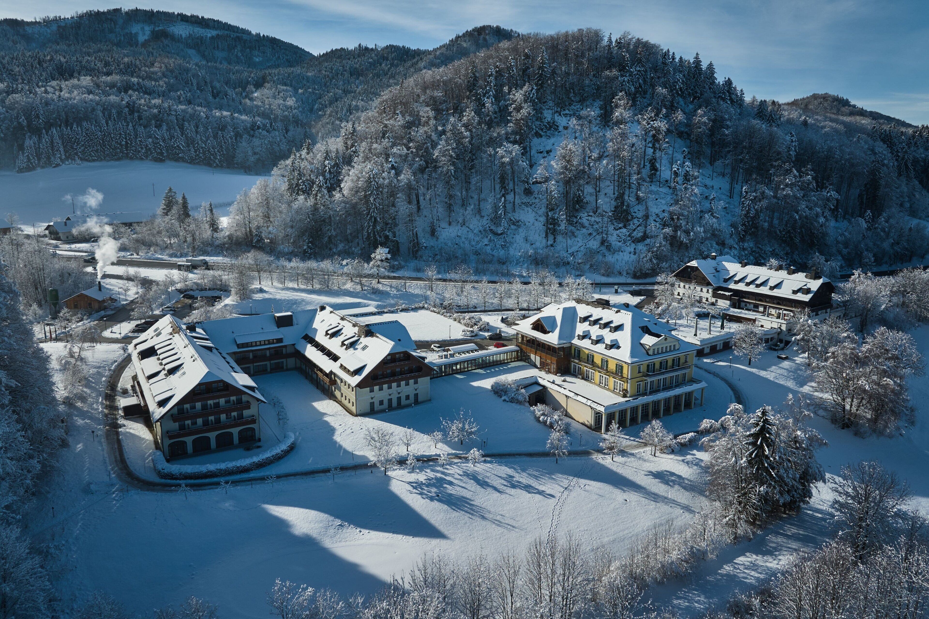 Un complejo turístico cubierto de nieve con varios edificios, incluyendo un gran edificio principal y cabañas más pequeñas, rodeado de un área montañosa boscosa.