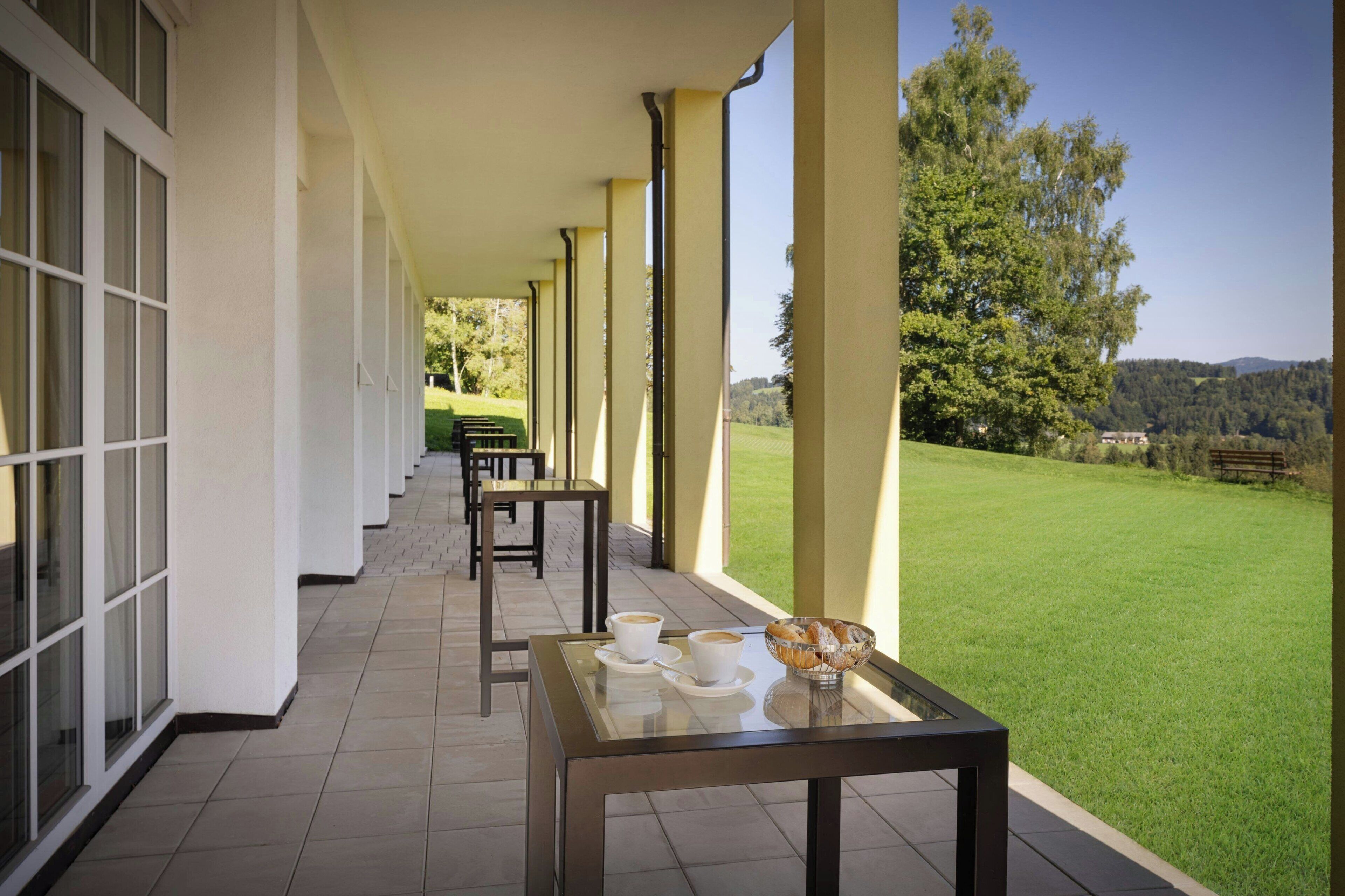 A covered outdoor area with tables and chairs, a glass-top table with cups and a bowl, and a view of a green lawn and trees.
