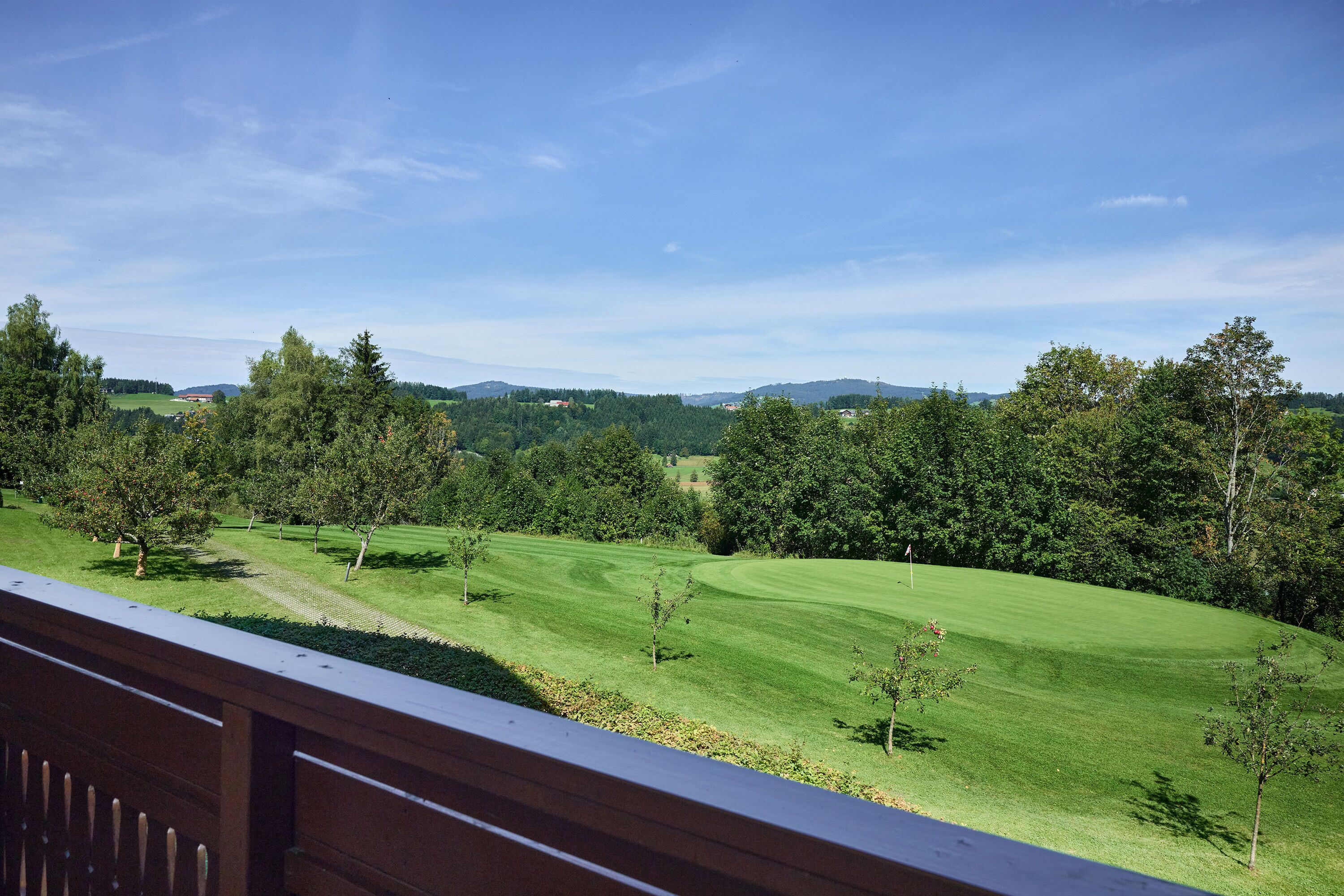 A view from a balcony overlooking a well-manicured golf course with trees and distant hills.