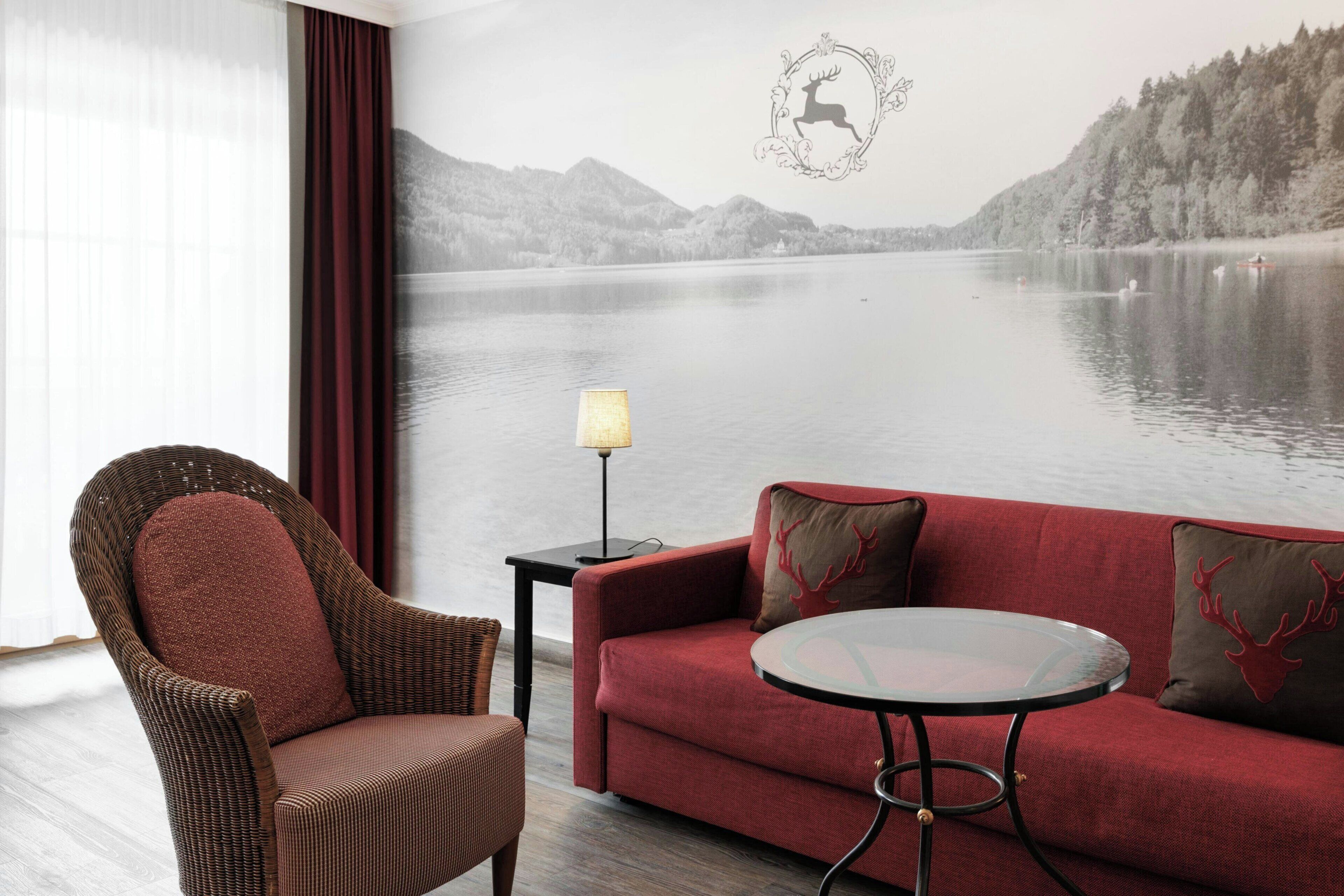 Habitación con vista a un lago, una silla de mimbre, un sofá rojo y una pequeña mesa con una lámpara.