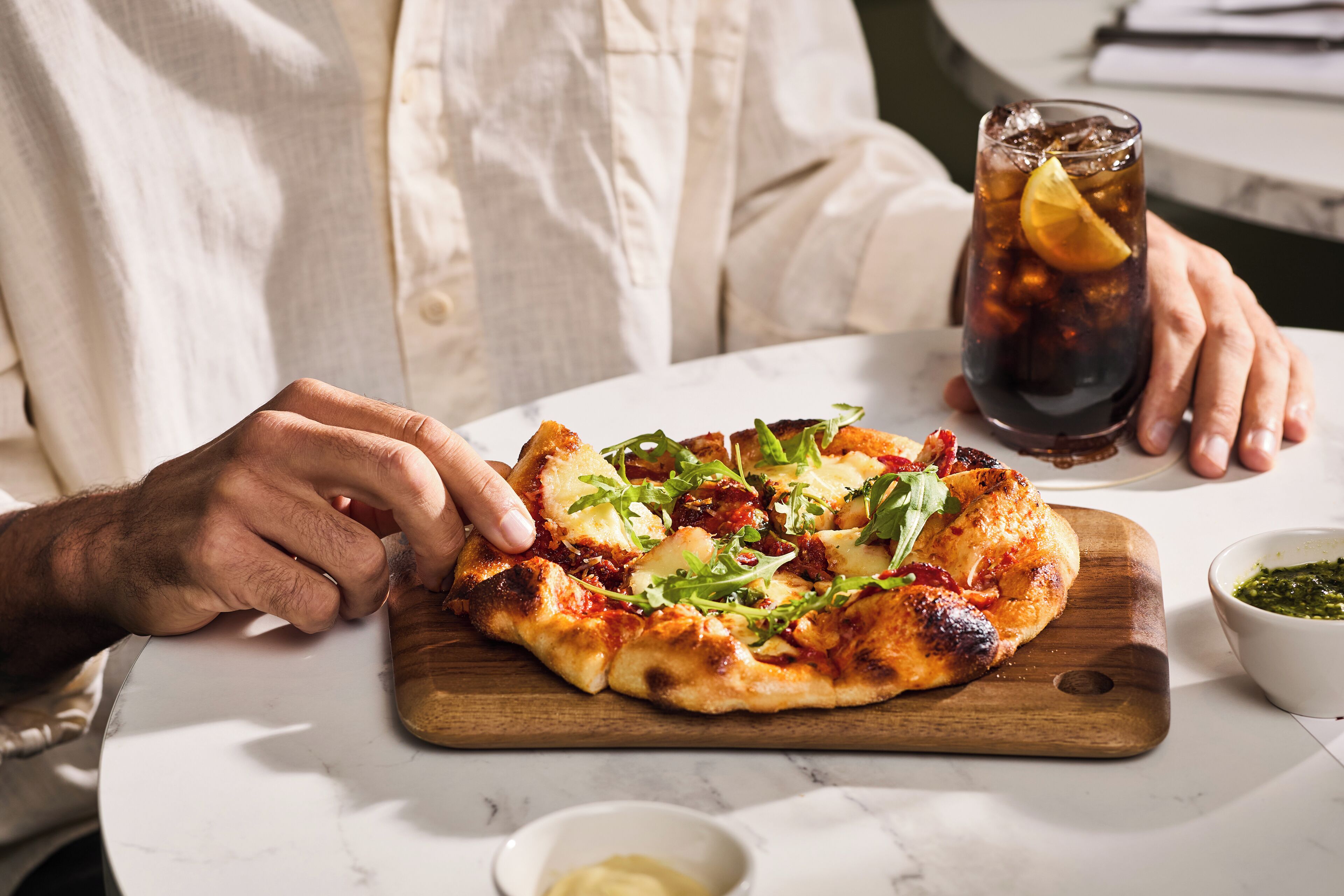 A person holding a slice of pizza on a wooden board, with a glass of iced beverage and a small bowl of sauce on the table.